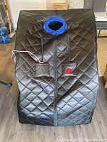 Front view of black quilted sauna tent with zipped front opening, Therasage logo and neck opening ring.