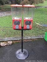 Full view of the vintage double gumball machine featuring two clear plastic hoppers with orange-red lids on a black metal stand outdoors.