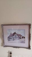 Frontal view of original artwork framed, showing a quaint small business building, possibly a hotel. Detailed depiction in soft colors, with visible signature (illegible) at bottom right corner.