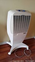 Front angled view of white cooling fan on a wheeled base, showing the protective grille and cord on floor.