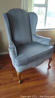 Front angled view of the gray fabric highback wingback chair with wooden cabriole legs on a wood floor near window.