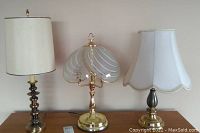 Photo showing three lamps on a wooden surface, varying designs and shades, includes a lamp with a glass shade, a lamp with a simple white fabric shade, and a lamp with a white scalloped fabric shade partially visible.