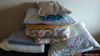 Overview of all linens including pillows, throw, mattress pad, and covers stacked on a floor against a white wall.