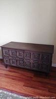 Front view of mid century modern wooden storage chest with raised geometric front panel design, dark finish, standing on hardwood floor.