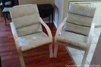 Pair of outdoor chairs with white frames and light patterned cushions showing weathering and stains, placed indoors on wooden floor with background furniture.