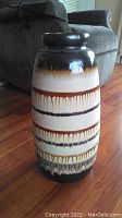 Full view of the West German pottery vase standing on floor near couch, showing vertical cylindrical shape with banded drip glaze in earth tones.