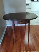 Front view of dark wood folding table showing oval top and drawer with metal pull.