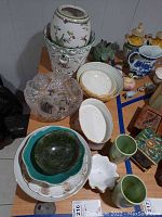 Wide view of ceramic bowls, platters, cups, and decorative items grouped on table showing variety of shapes, colors, and materials mainly ceramic with some glass.