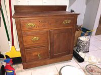 Solid wood cabinet with decorative carvings and brass hardware. Scratches and stains visible. Located in basement area with tiled floor and cleaning supplies nearby.