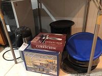 Photo showing boxed chafing dish, boxed wine corkscrew, black coffee thermos, black cast iron pan, roasting pan, and stack of plastic storage bowls with blue lid placed on a floor