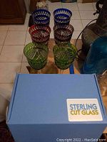 Top view of six colored pressed glass goblets in blue, red, and green arranged in two rows, and blue box labelled 'Sterling Cut Glass'.