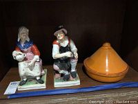Photo showing two porcelain figurines and a covered pottery bowl on a wooden shelf.