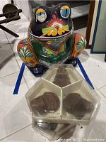 Front view of the colorful ceramic frog planter and the dragonfly yard ornament in a clear hexagonal container.