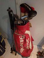 Photo showing red and white Spalding golf bag filled with assorted golf clubs including irons, drivers, and putters. Some clubs have head covers.