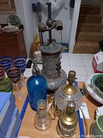 Photo showing two clear pressed glass lamps with brass fittings, a large samovar lamp conversion with colorful floral enamel, blue vintage seltzer bottle, and clear glass cruet on table