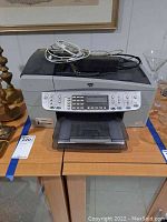 Front view of HP OfficeJet 6310 All-in-One printer on table showing cables on top and power cord behind.