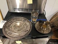 Full lot: two ornate plates, glass canister with lid and ladle, two cups, shot glass on black surface