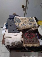 Photo showing a variety of linens and fabrics stacked on a table including a printed fabric with western horse and cowboy scenes, white linen, gray fabric with black squares, and a small fringed rug with a geometric design.