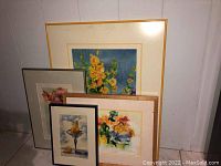 Four framed wall art pieces leaning against wall on tiled floor. Various floral and still life themes.