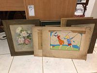 Framed floral painting and several unframed illustrations resting against wood panel wall.