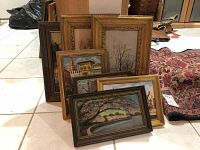 Seven framed paintings leaning on tiled floor, showing various landscapes and architectural scenes with different styles of frames.