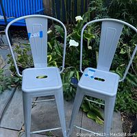 Two metal bistro bar stools placed outdoors on a patio. Both stools have a light grey finish and show a small blue sticker on the seats.