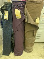 Photo of three nearly full bolts of upholstery fabric standing upright; fabrics include a blue paisley pattern bolt, a maroon striped bolt, and a beige ribbed bolt.