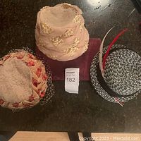 All three vintage hats aligned side by side on a dark counter: pink mesh with satin flowers, beige with red ribbon and netting, black and white woven with feathers