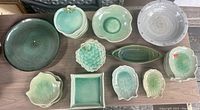 Top-down view showing various handmade ceramic dishes including fish, grape, vegetable shapes, and bowls in light green and grayish tones.