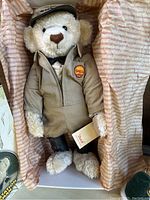 Teddy bear dressed in beige mechanic jumpsuit with Shell patch and cap, holding a tag, displayed inside the box with pink striped paper