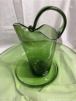 Green handblown glass pitcher with ruffled top and curved handle placed inside large green glass serving bowl
