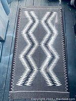 Full view of the rectangular Navajo wool rug laid flat, showing its geometric zigzag pattern in white, black and brown hues. Edges show minimal fraying and some wear.