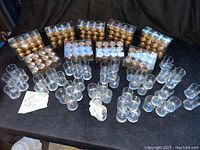 Photo showing 52 clear glass votive candle holders arranged in groups alongside 9 boxes of new gold votive candles in plastic packaging.