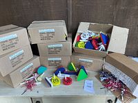 Photo showing multiple boxes filled with assorted racket raisen noisemakers and bundles of party horns displayed on table.