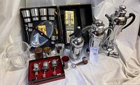 Vintage bar tools and accessories including crystal ice bucket, cocktail shakers, shot glasses, and various metal tools displayed on cloth