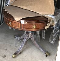 View of the round mahogany card table partially covered with cardboard showing the polished wood surface, drawer front with brass ring pull knob, and part of the pedestal base.
