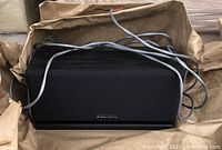 Front view of black Atlantic Technology home theater speaker with fabric grille and attached cables