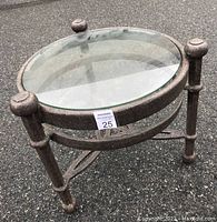 Cast aluminum patio end table with round tempered glass top shown outdoors on asphalt surface. Grey textured finish, three legs connected with braces, decorative finials visible.
