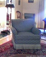 Green and gold patterned brocade rotating chair shown from the front in a living room setting with a fireplace nearby.