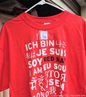 Close-up of one red T-shirt hanging on hanger showing multilingual 'I AM' text and 'RED HAT' in center.