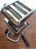 Full view of the chrome pasta machine with removable crank handle and clamp placed next to it on a wooden surface.