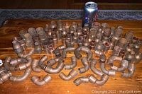 Brass plumbing parts including straight pieces, elbows, end cap, and outdoor faucet arranged on wooden surface with soda can for scale.