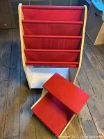 Children's book rack with red fabric shelves standing upright next to matching red-padded stepstool on wooden floor.