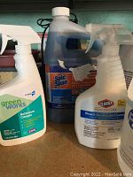 Close-up of various spray bottles including Clorox Healthcare Bleach Germicidal Cleaner and Green Works Bathroom Cleaner with Spic and Span gallon jug in background.