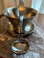 Photo of the stainless steel sauce bowl with ladle in front of original box, showing bowl, ladle, and some plastic wrapping.