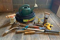 Full lot view showing watering can, black garden edging, pruning shears, bulb planter, dial spray bottle, weed puller and other garden hand tools scattered on wooden floor