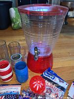 Red and clear plastic drink dispenser with tap, two clear glass beer mugs, patriotic mason jar style containers, tea light holders, and star-shaped confetti