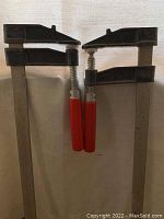 Front view of two large metal clamps positioned side by side showing red handles and black clamping heads.