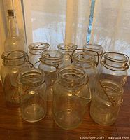 Ten vintage Ball canning jars with wire bail closures in clear glass and a tall antique glass bottle with a cork stopper arranged on a table under natural light.