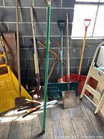 Wide view of assorted garden and yard tools leaning against cinder block wall including wooden handled shovels, shovels, manual garden tools, and two-man saw.